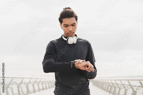 Young african-american mixed-race male athlete sportsman checking time on his wrist watch in fitness clothes preparing for jogging running on a city bridge in urban area. Active lifestyle concept