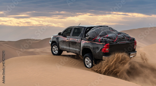 a black pickup truck is going up from a sand dune and splashing sands on air and around in dasht e lut or sahara desert with cloudy sky