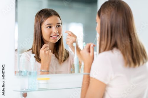 dental care, hygiene and people concept - happy smiling teenage girl with floss cleaning teeth and looking in mirror at bathroom