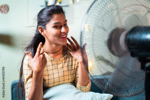 Happy smiling Indian Woman enjoying fan air during to heat stroke while sitting on couch - concept of heat wave or feeling cool air from fan at home.