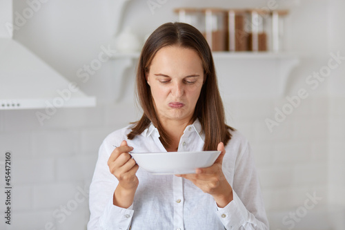 Dark haired Caucasian woman in white shirt demonstrates disgust twisting face with negative reaction while trying to eat some smelly soup from a plate in kitchen.