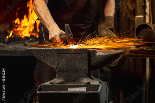 Close-up working powerful hands of male blacksmith forge an iron product in a blacksmith. Hammer, red hot metal and anvil. Concept of labor, retro professions