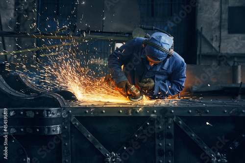 Competent industrial worker dressed in protective clothes, glasses and gloves grinding metal construction using polishing machine. Sparks from metal processing.