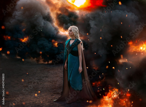 Art photo. Fantasy woman queen, blonde hair in braids. Warrior princess girl stands on background of black smoke, burning fire, flame war concept. Medieval costume, dress, cloak cape, leather armor.