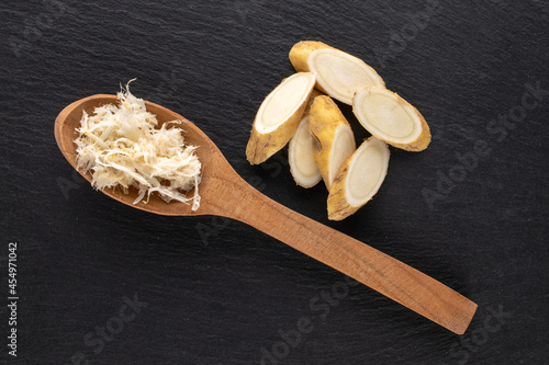 One cut into slices and grated horseradish roots with a wooden spoon on a slate stone, close-up, top view.