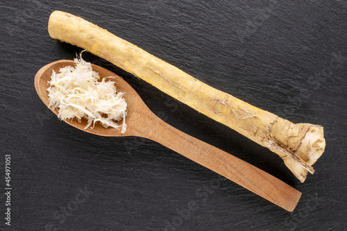 One whole and grated horseradish roots with a wooden spoon on a slate stone, close-up, top view.
