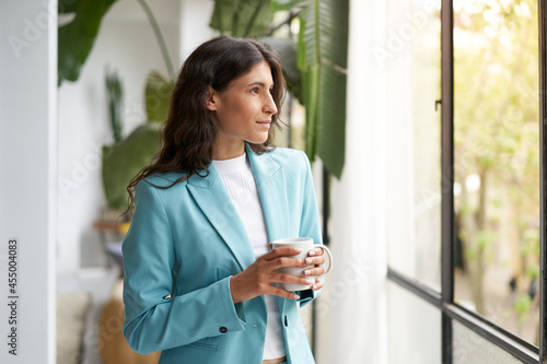 Young business woman satisfied with a job well done relaxing with her morning coffee or tea, looking out the window. Beautiful latina woman celebrating successes on break from work at the office.