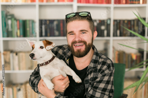 Happy male owner of jack russell terrier dog, feels responsibility of caring about pet, standing against bookshelves background. People and relationship with animals