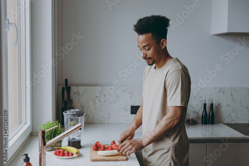 Side view young bachelor single african american man 20s wearing casual clothes prepare sweet salad chop cut fruit cooking food in light indoor kitchen at home alone. Healthy diet lifestyle concept