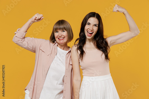 Two young strong sporty fitness daughter mother together couple women in casual beige clothes show biceps muscles on hand demonstrating strength power isolated on plain yellow color background studio.