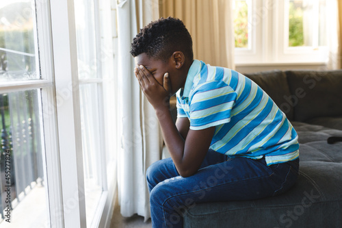 Sad african american boy covering his face and sitting at window in living room