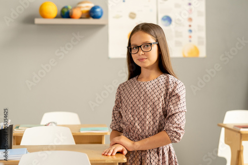 Attractive schoolgirl in dress and eyeglasses standing by her desk against pictures and models of planets
