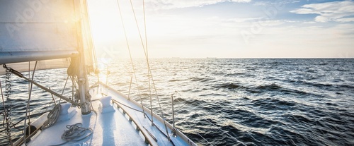 Yacht sailing in an open sea at sunset. Close-up view of the deck, mast and sails. Clear sky after the rain, glowing clouds, golden sunlight. Panoramic seascape