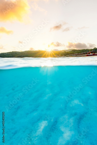 (Selective focus) Split shot, over under picture. Crystal clear, turquoise water and some beach umbrellas illuminated during a stunning sunrise. Grande Pevero Beach, Sardinia, Italy.