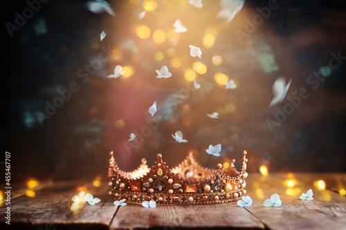 low key image of beautiful queen or king crown over wooden old table and falling flowers. fantasy medieval period