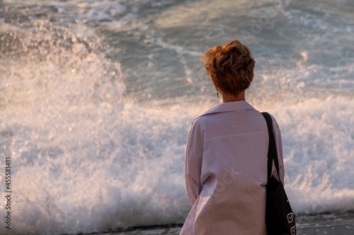 Rear view of caucasian woman with short hair hairstyle standing on city embankment and looking at large waves of stormy sea at sunset. Selective focus. Copy space. The theme of human and nature.