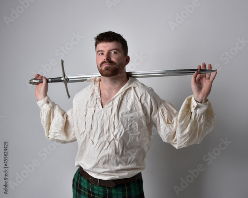 Close up portrait of handsome brunette man wearing Scottish kilt and renaissance white pirate blouse shirt. Holding a sword weapon, action pose isolated against studio background. 