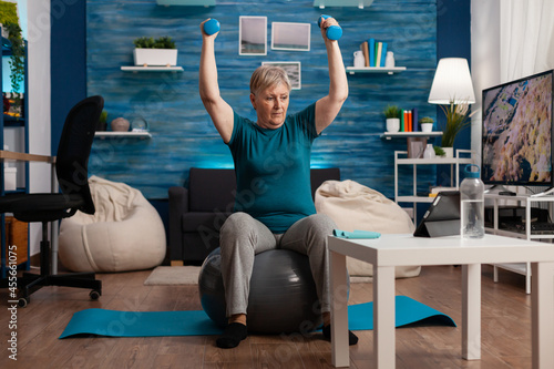 Focused retirement pensioner sitting on fitness swiss ball raising hand streching arm muscle doing healthcare exercise. Senior woman exercising body resistance using dumbbells in living room