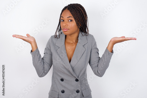 Puzzled and clueless Young african american business woman with braids over white wall with arms out, shrugging shoulders, saying: who cares, so what, I don't know.