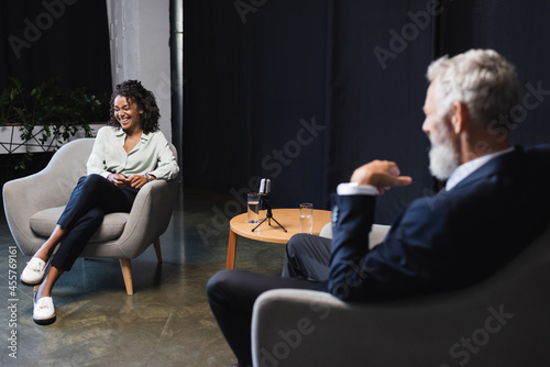 happy african american journalist laughing near blurred businessman during talk show