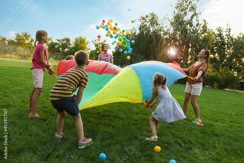 Group of children and teachers playing with rainbow playground parachute on green grass. Summer camp activity