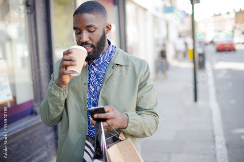 Young man with coffee and shopping bags texting with cell phone on urban sidewalk