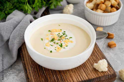 Homemade cauliflower soup puree in a white bowl with cheese, cream, spices and fresh parsley on a gray concrete background. Top view, copy space.