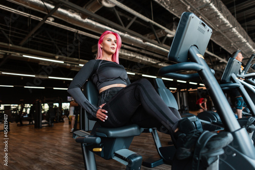 Sporty beautiful young fitness model woman with pink hair in fashionable black sportswear sits and trains on a stationary bike in the gym