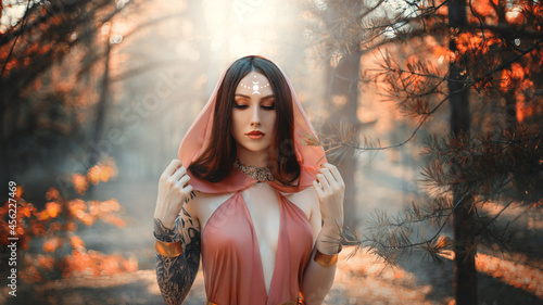 Fantasy woman priestess pagan witch, moon sign on forehead. Lady elf in autumn forest. Dark trees background, fog park. Girl princess in orange silk dress, hood on head. Sexy red haired fashion model.