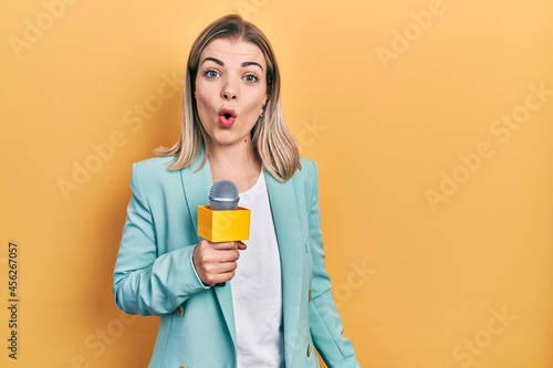 Beautiful caucasian woman holding reporter microphone scared and amazed with open mouth for surprise, disbelief face