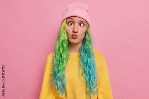 Crazy female student with colored hair has fun alone awkward expression foolishes around after all day ofstudying crosses eyes pouts lips wears hat and jumper poses against pink studio background.