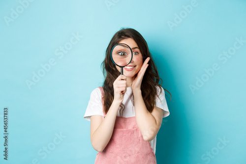 Girl searching for you. Cute smiling woman recruiter look through magnifying glass, staring at camera, investigating, looking for someone, standing over blue background