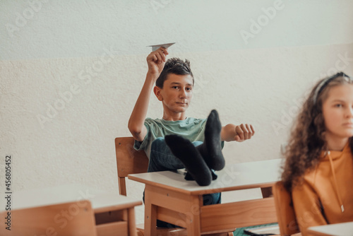 A naughty boy with his feet on a desk sits in the classroom and throws a plane. Selective focus