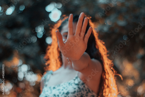 Mujer joven de pelo rizado evitando una fotografia haciendo la señal de parar o stop junto a la naturaleza de los arboles y la puesta de sol por la mañana