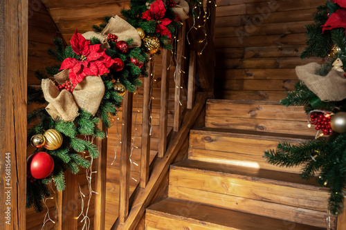 Detail house wooden staricase handrails railings decorated with artificial holly poinsettia flower, burlap bow, christmas tree and golden lights garland. Xmas family home interior decor idea concept