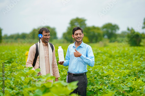 Young indian agronomist giving liquid fertilizer bottle to farmer and saying product information at green agriculture field.