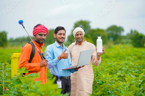 Young indian agronomist giving liquid fertilizer bottle to farmer and showing product information in laptop at green agriculture field.