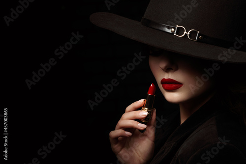 Beauty portrait of elegant woman with luxury lips makeup holding red lipstick. Model hides her face under wide hat, posing on black background. Close up studio portrait. Copy, empty space for text