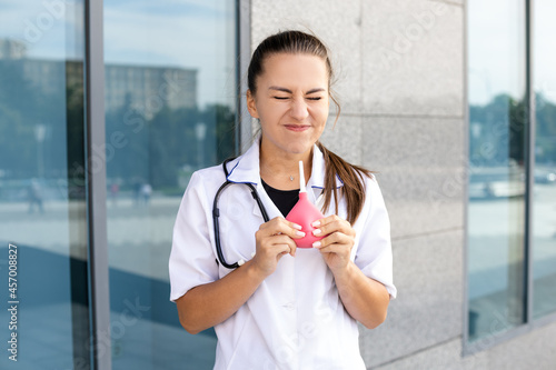 Medicine, healthcare, profession and people concept - A funny European woman doctor in a white coat with a stethoscope blows air on herself from an enema she holds in her hands outside