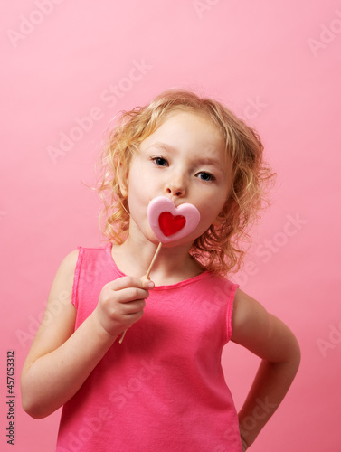 Little girl holding large heart-shaped lollipops in her hands on a pink background.