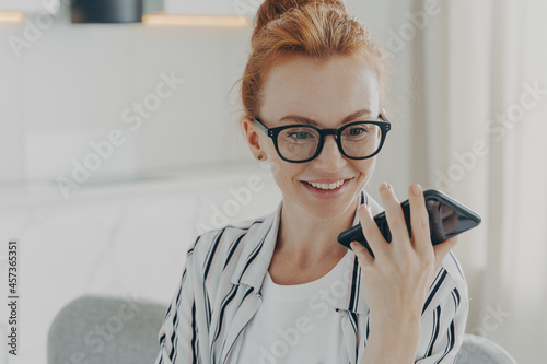 Beautiful positive red-haired woman in spectacles holding mobile phone and sending audio message