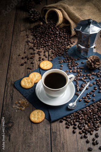 Hot coffee in a white coffee cup and many coffee beans placed around and sugar on a wooden table in a warm, light atmosphere, on dark background.