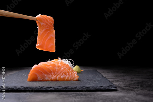 The hands were holding the chopsticks to hold the salmon sashimi, which was arranged on a black stone plate on an old table, with copy space.