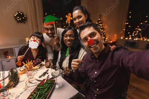 holidays, friendship and celebration concept - multiethnic group of happy friends with party props having christmas dinner at home and taking selfie