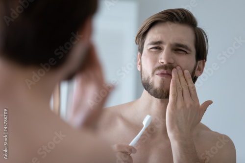 Handsome young man look in mirror touch cheek with hand, feels pain while brushing teeth due cavity or caries. Toothache, dental check up, oral care, need toothpaste for sensitive tooth enamel concept