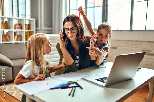 A business woman and a mother are trying to work on a laptop when her little daughters are playing, fooling around and interfering with her. Freelance, work from home.
