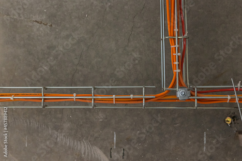 Cable tray with orange wires and metal duct show above t-bar grid below concrete of upper floor at construction site of building. Fire extinguishing and fire alarm system orange wires