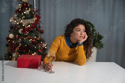 Young sad woman sits alone in an apartment in front of a Christmas tree and drinks champagne because she is alone for the first time for the holidays while her husband or fiance is a soldier in army
