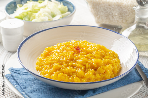 Italian dinner with risotto alla milanese and fresh salad. Italian dish made from saffron, rice, butter, hard cheese and vegetable broth. Raw arborio rice on background. White table.