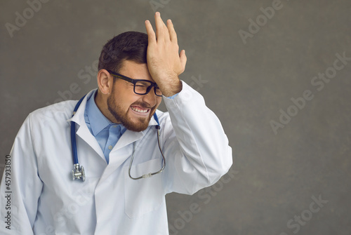 Damn idiot. Emotional male doctor smacks forehead as he remembers stupid mistake, foolish medical fiasco or professional failure at hospital, feeling dumb, annoyed and awkward, copy space background